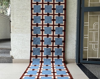 May include: A long rectangular runner rug featuring a repeating grid pattern. The design consists of light blue squares centred within white squares, separated by dark brown horizontal and vertical lines that create a structured geometric layout.