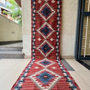 May include: A long, rectangular red rug with a geometric pattern. The rug features diamond shapes in blue, white, and grey, with a fringe at the bottom. The rug is approximately 244 cm long.