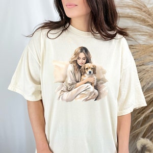 May include: A cream-colored t-shirt with a graphic print of a woman holding a dog. The woman and dog are both brown and white.