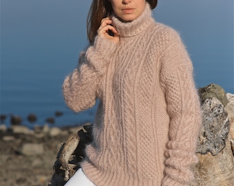 Può includere: Un maglione morbido e rosa, con collo alto e motivo a treccia. Il maglione è indossato da una persona in piedi davanti a uno specchio d'acqua e un grande pezzo di legno alla deriva.