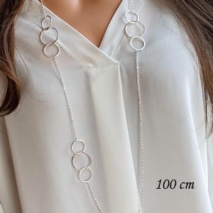 Op de afbeelding: Een lange zilverkleurige ketting met een fijne schakelketting afgewisseld met meerdere cirkelvormige ringen. De ketting heeft een totale lengte van 100 cm, zoals aangegeven in de tekst. Het ontwerp bestaat uit twee groepen van drie ringen verbonden door kettingsegmenten.