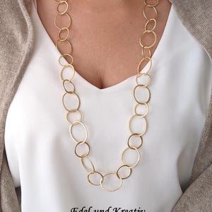 Puede incluir: Un collar de cadena de oro con grandes anillos ovalados unidos. El collar lo lleva una persona con una camisa blanca y un cárdigan marrón.