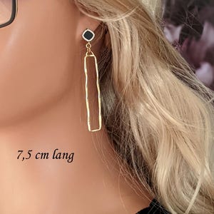 Op de afbeelding: Goudkleurige oorbellen met een zwart vierkant accent en een lange, rechthoekige hanger. De oorbellen zijn 7,5 cm lang. De tekst geeft de lengte aan.