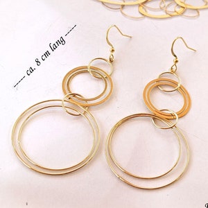 Könnte beinhalten: Goldfarbene Hängeohrringe mit einer Reihe von miteinander verbundenen Kreisen. Die Ohrringe haben einen Hakenverschluss und sind ca. 8 cm lang. Das Design umfasst mehrere Größen von kreisförmigen Formen, wodurch ein geschichteter Effekt entsteht.