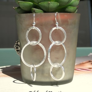 Könnte beinhalten: Silberne Ohrringe mit drei ineinandergreifenden Kreisen. Die Ohrringe sind silberfarben und haben eine strukturierte Oberfläche.