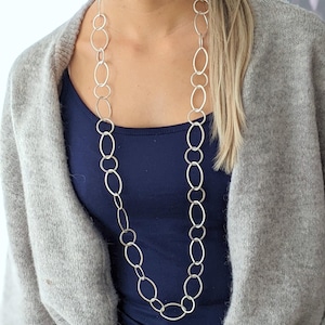 May include: A long silver chain necklace with oval and circular links. The necklace is worn over a navy blue top and a gray cardigan. The text "Edel-und-Kreativ" is visible in the upper right corner.