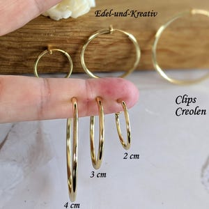 Könnte beinhalten: Eine Sammlung goldfarbener Creolen in verschiedenen Größen mit Durchmessern von 2 cm, 3 cm und 4 cm. Die Ohrringe sind mit Clips Creolen und Edel-und-Kreativ beschriftet. Diese kreisförmigen Ohrringe haben eine glatte, polierte Oberfläche.