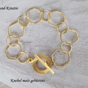 Puede incluir: Una pulsera de tono dorado que presenta una serie de eslabones circulares y hexagonales alternados con un acabado mate cepillado. La pieza se asegura con un cierre de palanca que consiste en un anillo circular y una barra. El texto Edel-und-Kreativ y Knebel matt gebürstet es visible.