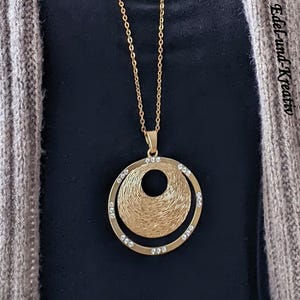 May include: Gold-toned pendant necklace featuring a double-circle design. The inner circle has a textured, woven appearance, while the outer circle is adorned with small, clear crystals. The necklace hangs on a delicate chain.