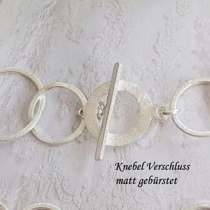 Könnte beinhalten: Nahaufnahme eines silberfarbenen Halskettenverschlusses mit Knebelverschluss. Der Verschluss ist mit einer Reihe von strukturierten, kreisförmigen Gliedern verbunden. Der Text "Knebel Verschluss matt gebürstet" ist sichtbar.