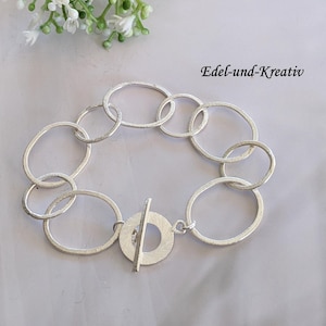 Könnte beinhalten: Ein silberfarbenes Armband mit einer Reihe von miteinander verbundenen ovalen und kreisförmigen Gliedern. Das Armband hat einen Knebelverschluss. Der Text "Edel-und-Kreativ" ist sichtbar.