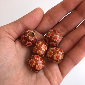 May include: Five wooden beads with a floral pattern in shades of brown, orange, and cream. The beads are held in a hand, showcasing their round and faceted shapes. One bead has a hole.