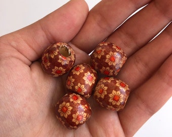 Puede incluir: Cinco cuentas de madera con un estampado floral en tonos marrón, naranja y crema. Las cuentas se sostienen en una mano, mostrando sus formas redondas y facetadas. Una cuenta tiene un agujero.