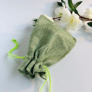 May include: A small, sage green drawstring bag made of woven fabric. The bag is gathered at the top with a bright green drawstring. White flowers are in the background.
