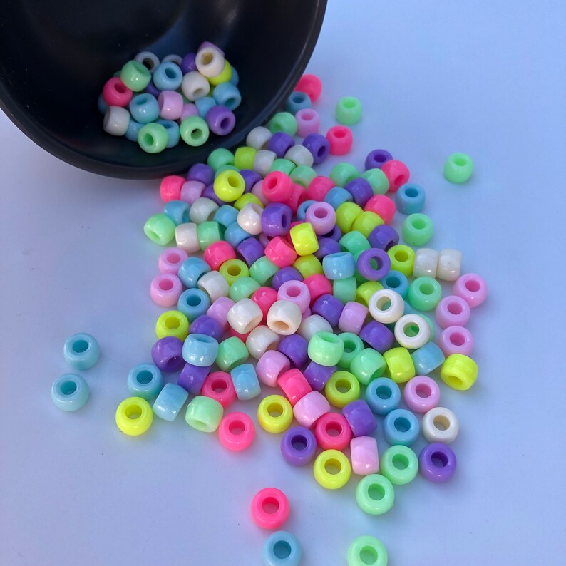 Puede incluir: Una colecci&oacute;n de cuentas de pony de colores pastel, incluyendo rosa, amarillo, verde, morado y azul, derram&aacute;ndose de un cuenco negro. Las cuentas son redondas con un agujero en el centro, ideales para proyectos de manualidades.