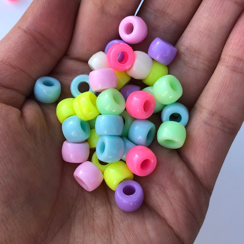 Puede incluir: Una colecci&oacute;n de cuentas de pl&aacute;stico redondas y coloridas en varios tonos pastel, incluyendo rosa, azul, amarillo y morado. Las cuentas tienen forma cil&iacute;ndrica con un agujero en el centro, adecuadas para ensartar. Las cuentas se muestran en una mano.