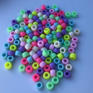 Puede incluir: Un mont&oacute;n de cuentas de pony de colores en varios tonos de rosa, morado, verde, amarillo y blanco. Las cuentas son cil&iacute;ndricas con un agujero grande en el centro, perfectas para ensartar. Las cuentas est&aacute;n esparcidas sobre una superficie azul claro.