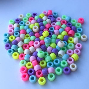 Puede incluir: Un conjunto de cuentas de pony de colores pastel, incluyendo rosa, morado, amarillo, verde y azul. Las cuentas son cil&iacute;ndricas con un agujero grande en el centro, adecuadas para ensartar. Las cuentas est&aacute;n dispersas sobre una superficie blanca.