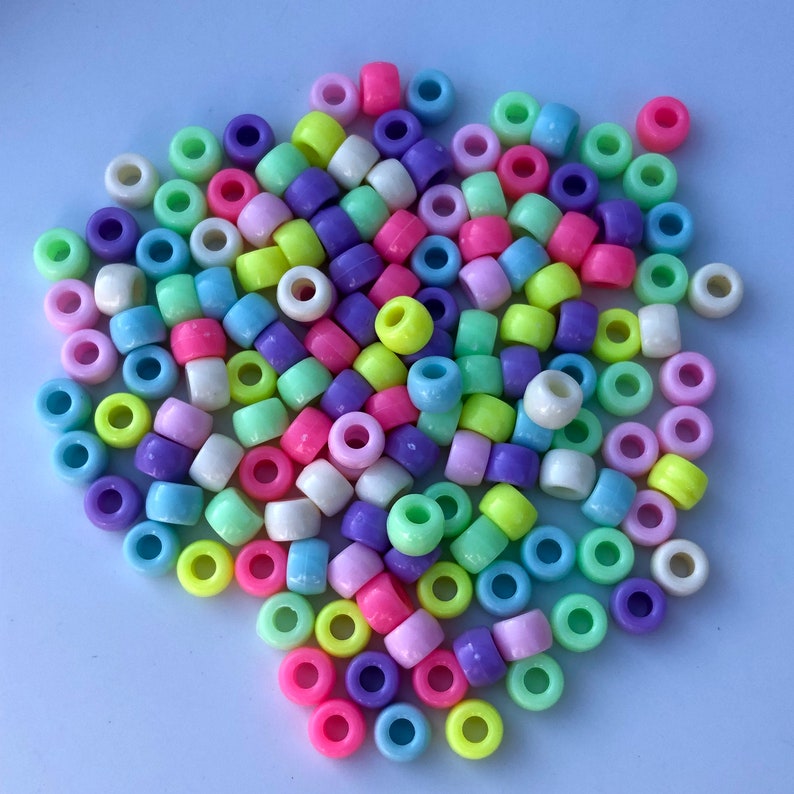 Puede incluir: Un mont&oacute;n de cuentas de pl&aacute;stico de colores en varios tonos de rosa, morado, verde, amarillo y blanco. Las cuentas son cil&iacute;ndricas con un agujero en el centro, adecuadas para la fabricaci&oacute;n de joyas o manualidades.