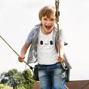 May include: A young boy with blonde hair is swinging on a rope swing. He is wearing a white t-shirt with a black smiley face on it, blue denim shorts, and a gray button-down shirt. He is smiling and laughing as he swings.