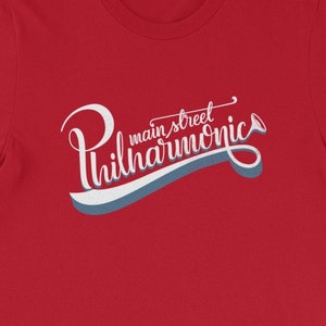 May include: Red short-sleeved t-shirt with a white graphic that says "main street philharmonics" in a stylized font.