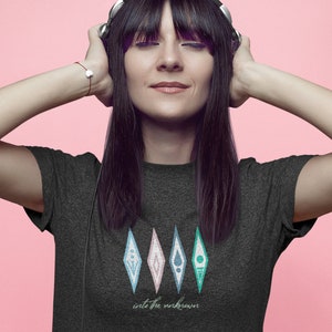 Puede incluir: Una mujer con el pelo largo y negro lleva una camiseta gris oscuro con un gr&aacute;fico de cinco diamantes de colores y el texto "into the unknown". Lleva auriculares y tiene los ojos cerrados.