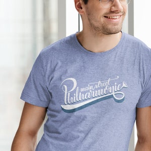 May include: A man wearing a light blue t-shirt with a white graphic that says "main street philharmonic" in a cursive font. The graphic also includes a small trumpet icon.