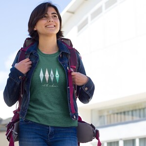 Puede incluir: Una mujer lleva una camiseta verde con un gr&aacute;fico de cuatro diamantes de color verde azulado y blanco con el texto "out for adventure" debajo. Lleva una chaqueta vaquera y una mochila.