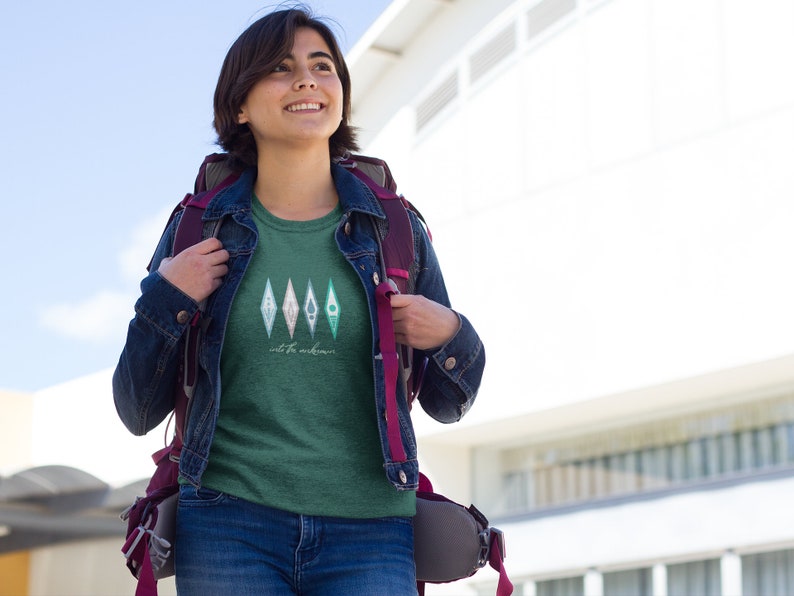 Puede incluir: Una mujer lleva una camiseta verde con un gr&aacute;fico de cuatro diamantes de color verde azulado y blanco con el texto "out for adventure" debajo. Lleva una chaqueta vaquera y una mochila.