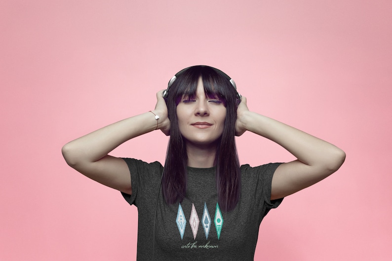 Puede incluir: Una mujer con el pelo largo y negro lleva una camiseta gris oscuro con un gr&aacute;fico de cinco diamantes de colores y el texto "into the unknown". Lleva auriculares y tiene los ojos cerrados.
