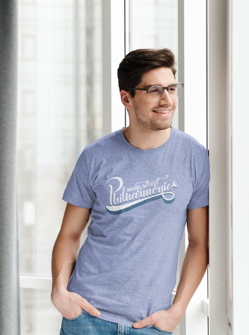 May include: A man wearing a light blue t-shirt with a white graphic that says "main street philharmonic" in a cursive font. The graphic also includes a small trumpet icon.