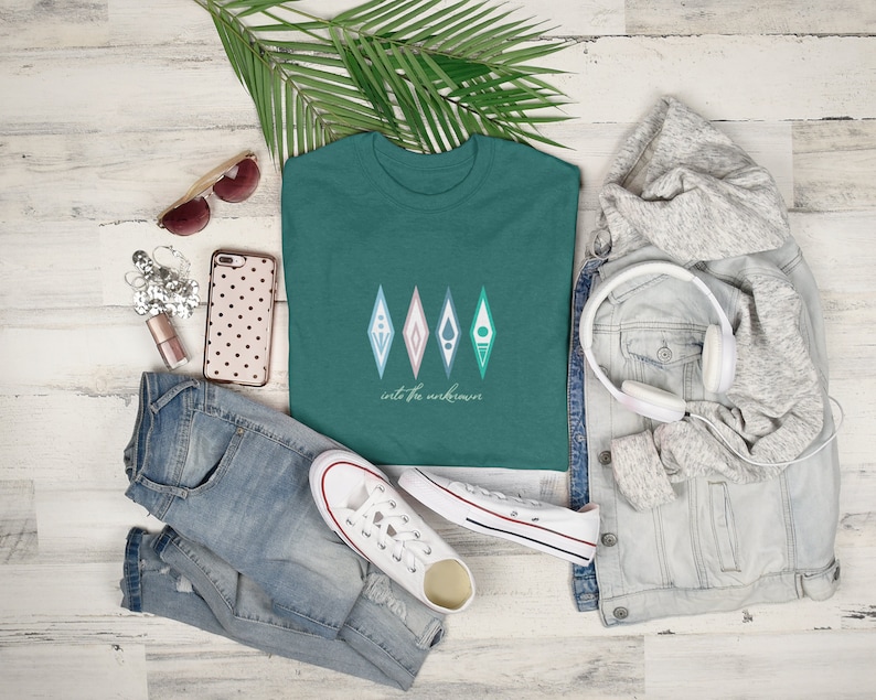 Puede incluir: Una camiseta verde azulada con un gr&aacute;fico de cuatro formas geom&eacute;tricas en tonos de rosa, azul y verde. El texto "into the unknown" est&aacute; impreso debajo de las formas.