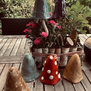Könnte beinhalten: Eine Sammlung von Keramikpilzskulpturen in verschiedenen Farben, darunter Blau, Rot mit weißen Punkten und Braun. Zwei hohe Pilze befinden sich in einem rechteckigen Pflanzgefäß aus Birkenrinde, das mit rosa Blumen und grünem Laub gefüllt ist.