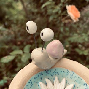 Könnte beinhalten: Eine Keramik-Schneckenfigur mit glänzend weißem Körper und blassrosa Gehäuse, ausgestattet mit zwei dünnen Metallstielen, die in weißen Augenperlen enden. Die Schnecke befindet sich auf einem runden, hellen Keramiksockel mit blau-weißem Blumenmuster.