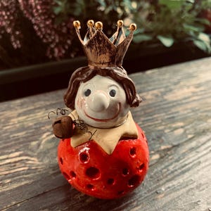 Può includere: Figurina in ceramica di un giullare incoronato. La figura ha un viso bianco, capelli castani e un corpo rosso a forma di fragola con piccoli fori. La corona è dorata e il giullare tiene una campanella.