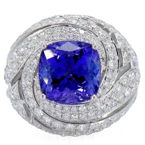 Op de afbeelding: Een oogverblindende ring met een grote, vierkant geslepen, diepblauwe edelsteen in het midden. De edelsteen is omgeven door een halo van kleinere, sprankelende witte diamanten, geplaatst in een wervelend, sierlijk ontwerp. De ringband is ook bezet met diamanten.