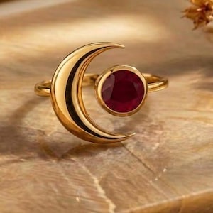 Könnte beinhalten: Ein goldener Ring mit einer Mondsichelform auf der einen Seite und einem runden, tiefroten Edelstein in einer kreisförmigen Fassung auf der anderen Seite. Das Metallband ist poliert und glatt und bildet ein offenes Design, das den Kontrast zwischen dem Gold und dem roten Stein hervorhebt.