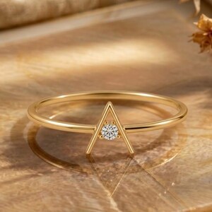 May include: A thin gold ring featuring a triangular geometric centrepiece. The centre of the triangle is set with a single, small, clear round-cut gemstone that sparkles under the light. The band is smooth, polished, and circular in shape.