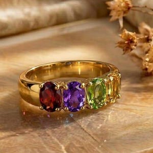 Op de afbeelding: Een gouden ring met vier ovaal geslepen edelstenen op een rij. De stenen bestaan uit een dieprode granaat, een levendige paarse amethist, een heldergroene peridoot en een gele citrien, gezet in een gepolijste geelgouden metalen zetting.