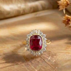 Puede incluir: Un anillo de oro con una gran piedra preciosa de color rojo intenso en talla cojín en el centro, rodeada por un halo de pequeñas piedras transparentes en talla marquesa. El aro es fino y metálico, con un diseño clásico y una pieza central facetada.