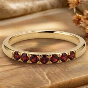 May include: A gold band ring featuring a row of seven round-cut deep red garnet gemstones set along the front. The metal has a polished finish, and the stones are arranged in a straight line, creating a consistent and symmetrical appearance.