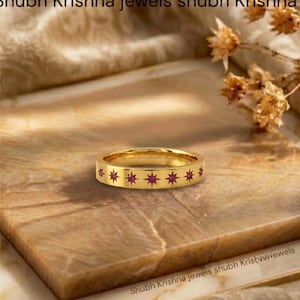 May include: A gold band ring featuring a repeating pattern of star-shaped engravings, each set with a small, round, vibrant pink gemstone. The ring has a polished metallic finish and a flat, uniform profile around the entire circumference.