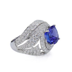 Op de afbeelding: Een oogverblindende ring met een grote, peervormige, diepblauwe edelsteen. De ringband is gemaakt van wit metaal en is bezet met talrijke kleine, sprankelende witte edelstenen, waardoor een halo-effect rond de centrale steen ontstaat. Elegant ontwerp.