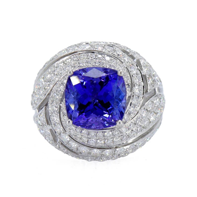 Op de afbeelding: Een oogverblindende ring met een grote, vierkant geslepen, diepblauwe edelsteen in het midden. De edelsteen is omgeven door een halo van kleinere, sprankelende witte diamanten, geplaatst in een wervelend, sierlijk ontwerp. De ringband is ook bezet met diamanten.