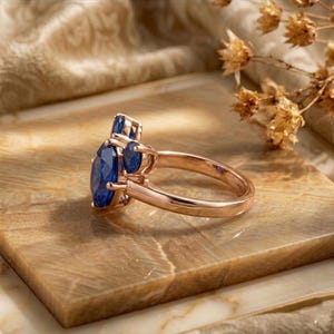 May include: A rose gold ring featuring a large oval-cut blue sapphire as the center stone, flanked by two smaller round-cut blue sapphires on each side. The band is smooth and polished, showcasing a classic three-stone design with prong settings.