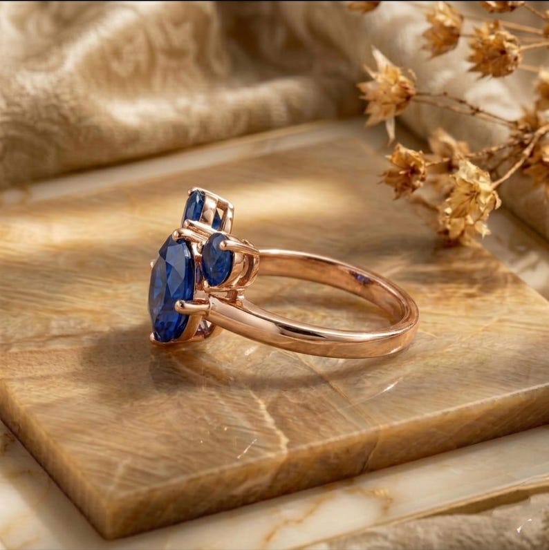 May include: A rose gold ring featuring a large oval-cut blue sapphire as the center stone, flanked by two smaller round-cut blue sapphires on each side. The band is smooth and polished, showcasing a classic three-stone design with prong settings.