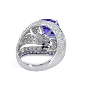 Op de afbeelding: Een zilveren ring met een grote, ovaal geslepen, diepblauwe edelsteen, omringd door meerdere rijen kleinere, heldere edelstenen. De ring heeft een brede, gedetailleerde band met een gepolijste afwerking.