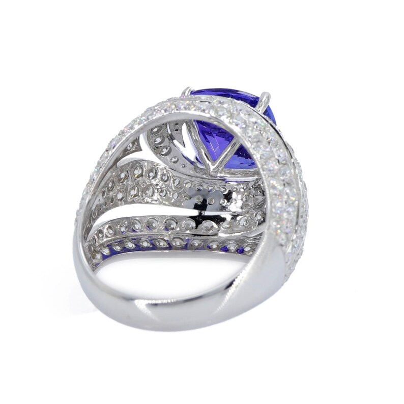 Op de afbeelding: Een zilveren ring met een grote, ovaal geslepen, diepblauwe edelsteen, omringd door meerdere rijen kleinere, heldere edelstenen. De ring heeft een brede, gedetailleerde band met een gepolijste afwerking.