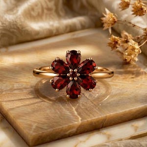 Può includere: Un anello in oro caratterizzato da un motivo floreale composto da sei granati rosso scuro a forma di pera, disposti attorno a un piccolo granato centrale. La fascia è sottile e lucida, con una tonalità metallica calda e brillante.