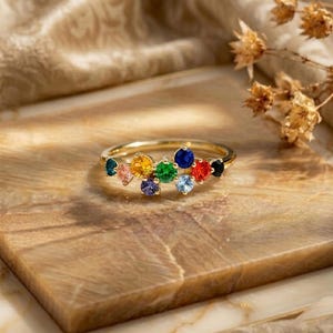 Può includere: Un anello in oro con un gruppo di nove pietre preziose a taglio rotondo di vari colori, tra cui blu, arancione, giallo, verde, rosso e nero. Le pietre sono disposte in una configurazione sfalsata su più livelli lungo la parte superiore dell'anello.